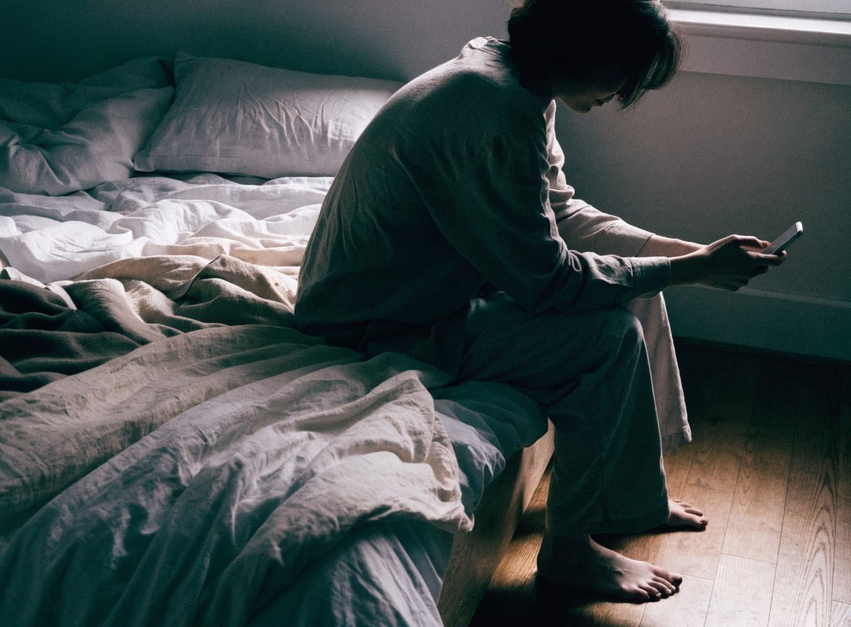 Person sitting on the edge of an unmade bed in early morning light, looking down at a phone