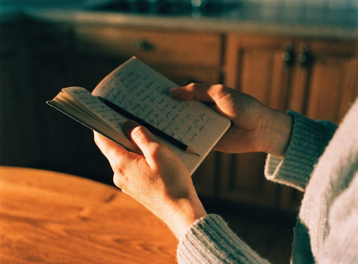 Close-up of hands holding an open notebook in a kitchen in warm evening light.