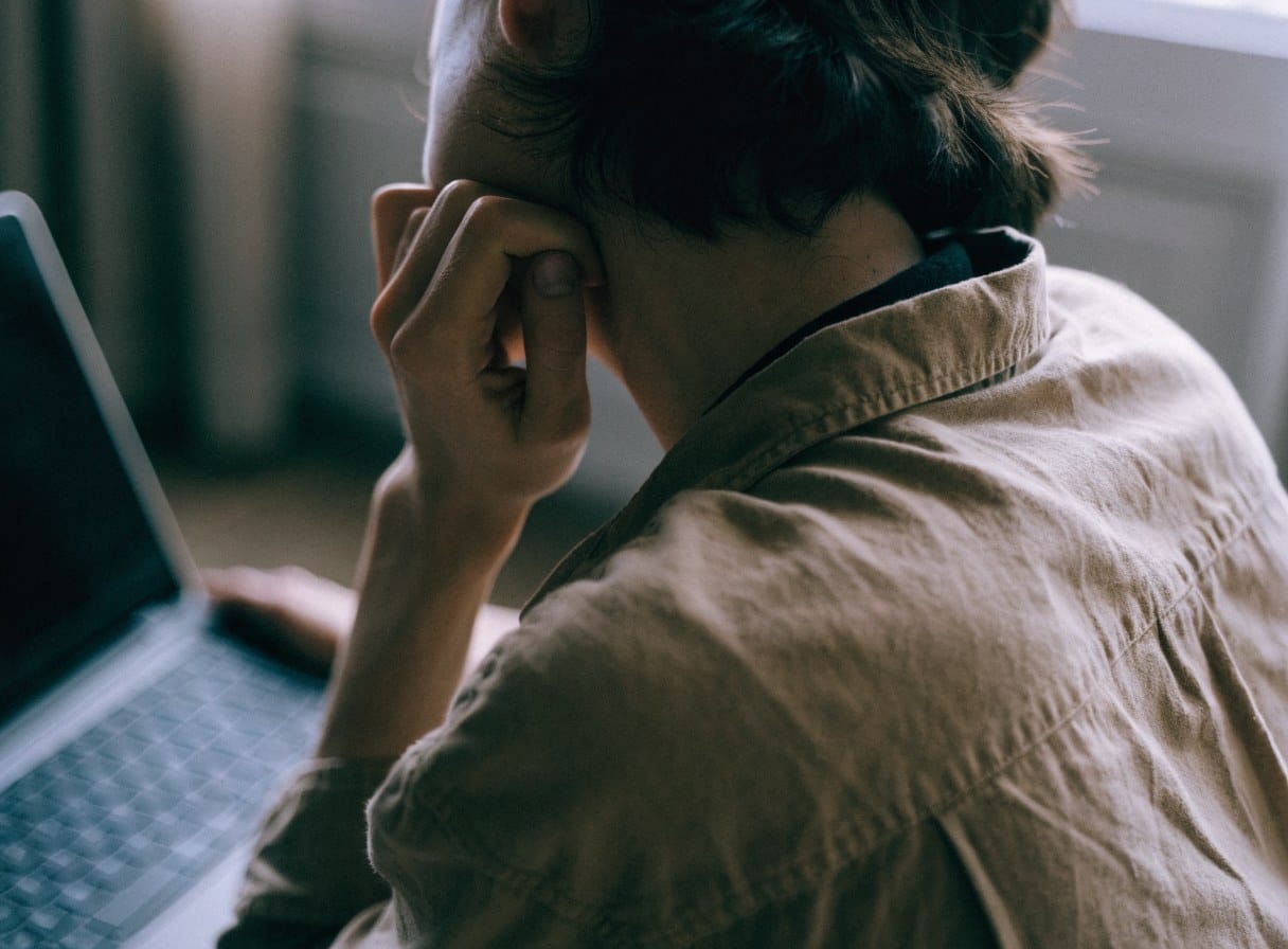 Close-up of a person at a laptop with shoulders held and a hand pressed to the side of their head, a sign of tension held at the desk.