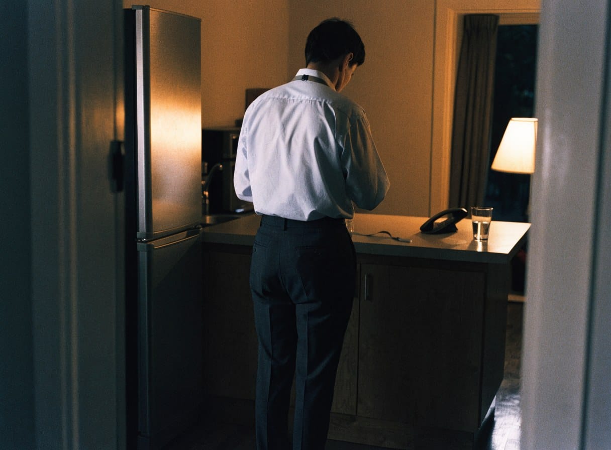 A person still in work shirt and trousers standing at a kitchen counter in the evening, a glass of water beside them under lamp light.
